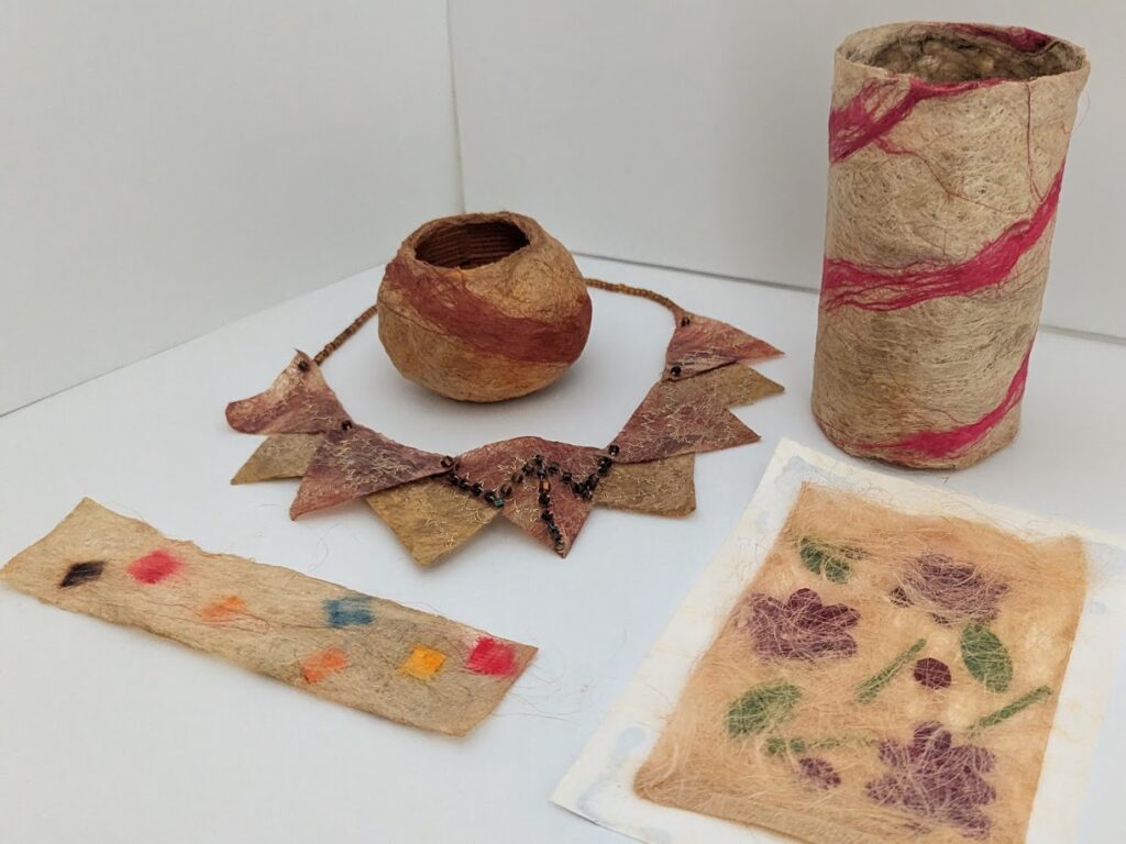 Felted flax samples, including a long thin rectangle of beige flax with squares of red, orange, yellow, blue and purple, a rectangle of beige flax with purple flowers and green leaves and stems embedded in it, a small bowl of beige with a diagonal red stripe, a necklace of purple and beige triangles, and a tall felt cylinder that is beige with a red stripe spiraling around it.