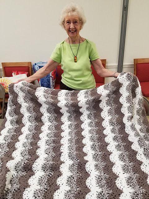 Jean Heckelmoser, a smiling elderly woman with white hair wearing a line green T-shirt holds up a crocheted blanket. The blanket is striped in a scallop pattern with a three stripe gradient from white to brown.