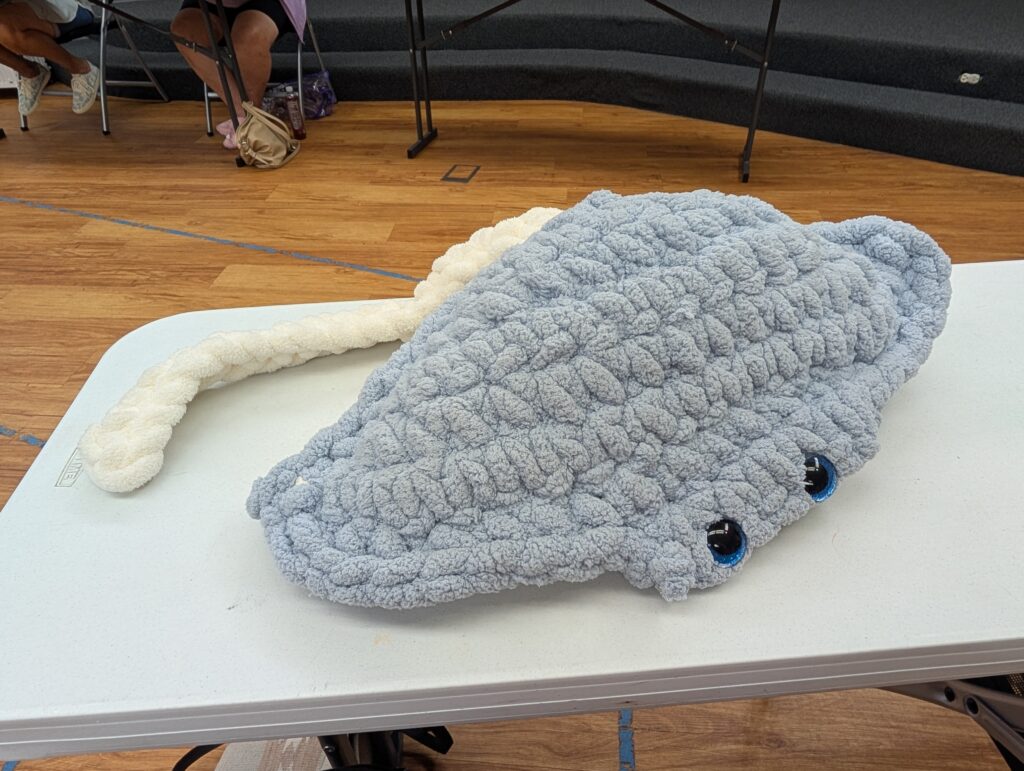 A large crochet plushie of a gray manta ray with a white tail.