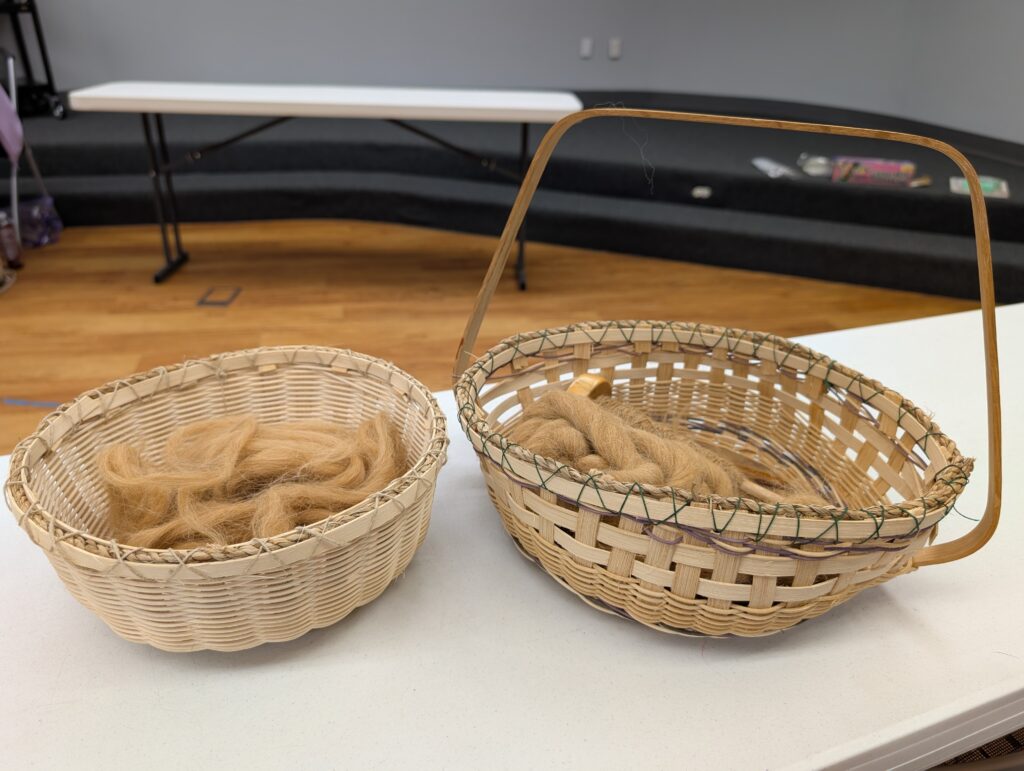 Two handwoven baskets. The left one is light tan woven of many thin reeds and has unspun fiber of a dark tan in it. The basket on the left is slightly darker, woven of larger slats. It has a handle and inside is more unspun fiber and a wooden top whorl spindle with a cop of singles.