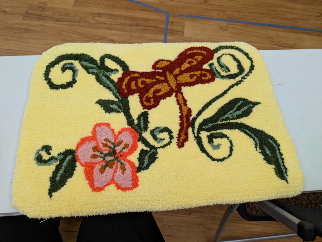 A tufted rug approximately the size of a bathmat. There is a pink flower, a red and yellow dragonfly, and a few green leaves on a field of yellow.