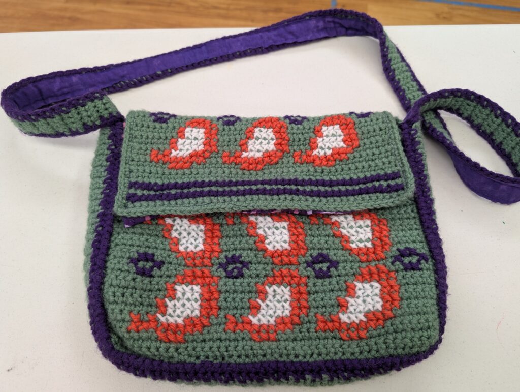 A small crochet purse in a drab green with dark blue trim. A leaf motif in orange and white has been cross stitched over the crochet.