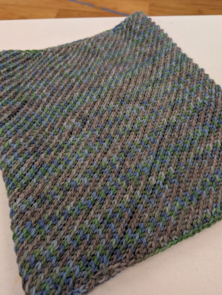 A knit cowl. The yarn is multicolored but overall appears as a mottle gray. The texture looks as if the knit stitches move diagonally across the cowl.