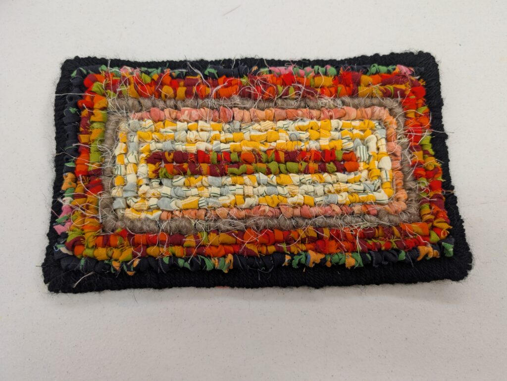 A small rectangular hooked rug in shades of orange, yellow, orange and black radiating outwards.