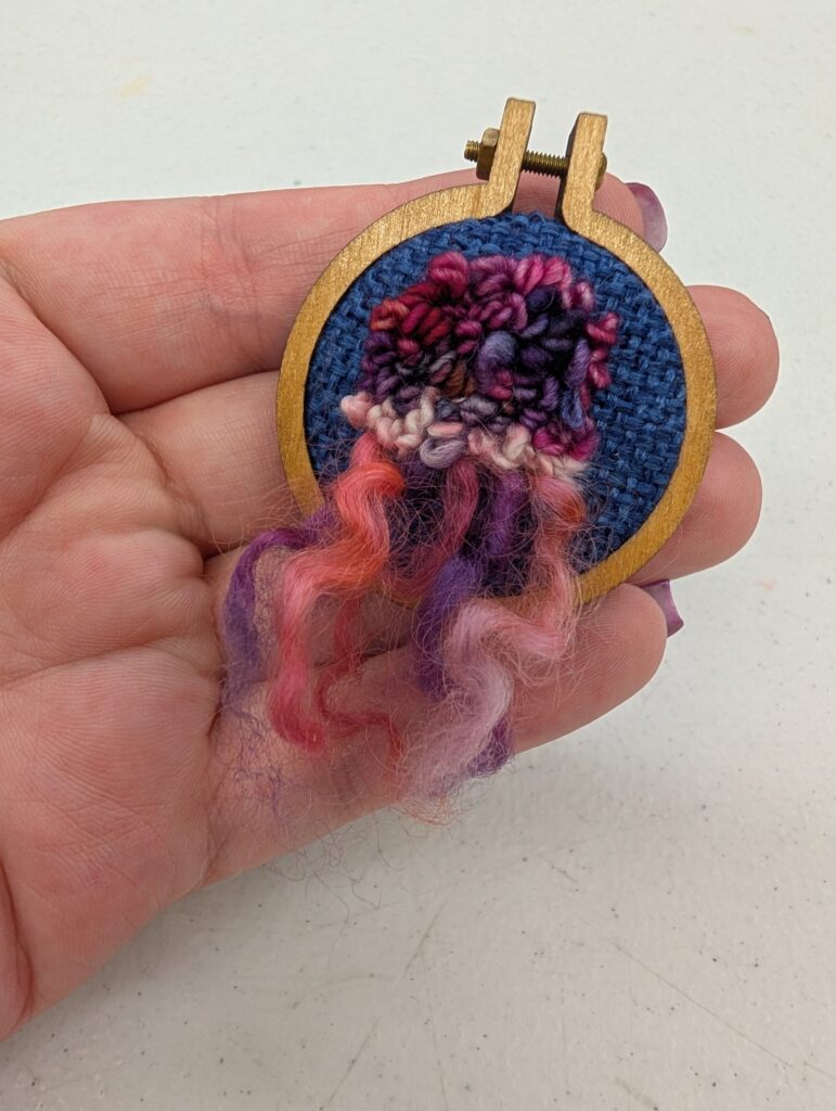 The tiniest embroidery hoop holding dark blue fabric. On the fabric is dark pink and purple yarn that has been rug hooked into it, the ends left long at the bottom to give the effect of a tiny jellyfish.