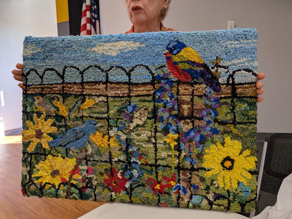 A rug hooked rug by Fonda Haddid. It features a garden scene. There is a large male painted bunting in the upper right corner, a black scalloped fence, large yellow flowers, re flowers, and small blue flowers, and three other birds.