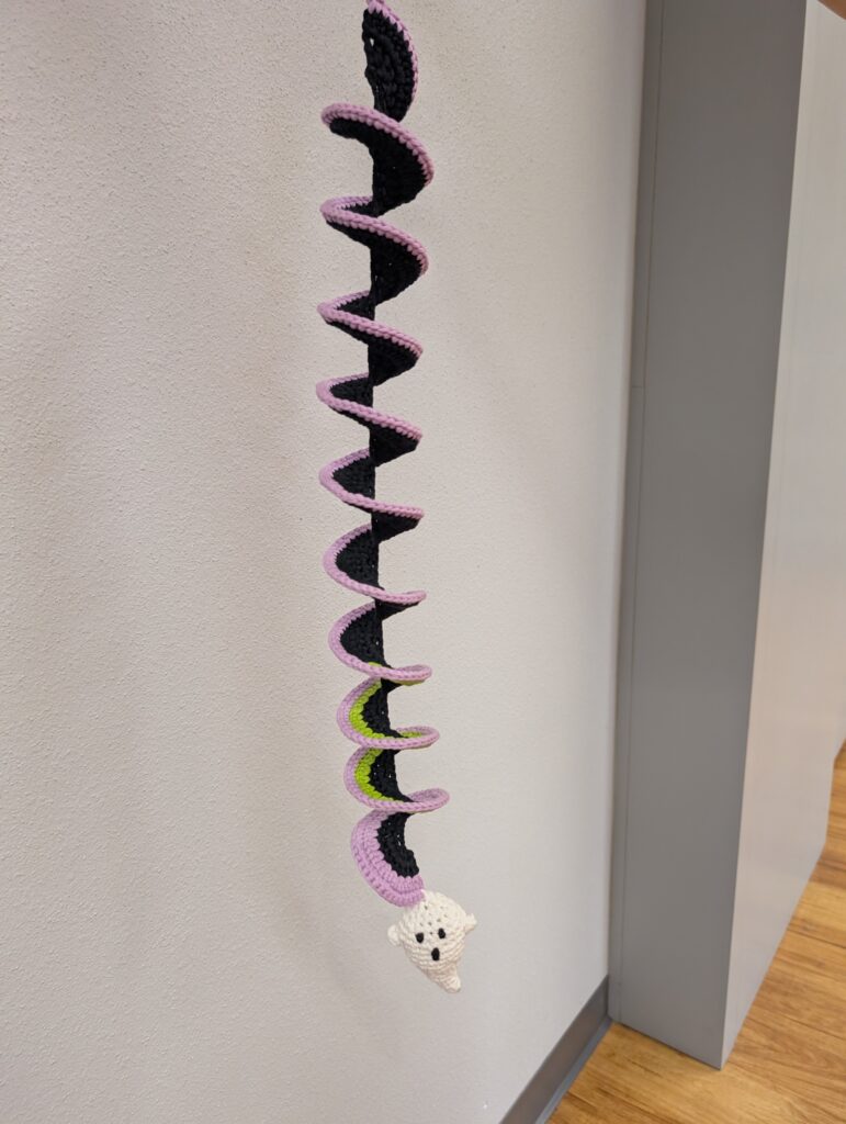 A long crochet spiral dangle. It is black with a purple edge and at the bottom is a little amigurumi ghost.