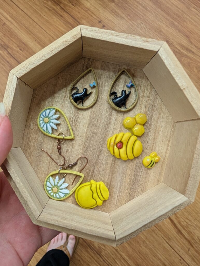 A wooden octagonal box corrals several small bits of polymer clay. There is one pair of finished earrings that are a yellow teardrop shape with a daisy in the round end. Two more gold teardrops have a black cat playing with a blue butterfly in them. There is also a yellow honey jar, a yellow beehive, a yellow bee, and a yellow three hexagon comb.