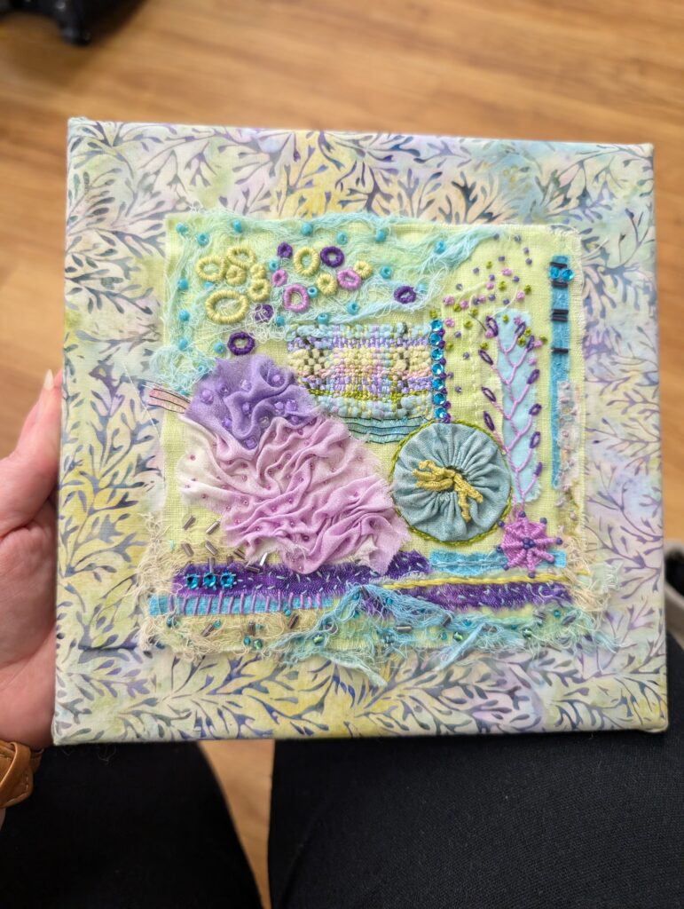 Mounted on a board covered with a pastel yellow and blue batik fabric, this embroidery features a number of different sashiko stitch techniques. It has a small weaving done on a mending loom, a blue fabric yo-yo, blue netting and beading, and a pink and purple fabric that's been sewn into a rippling shape. The overall colors are pastel yellow, blue, pink, and purple and gives the feeling of a coral reef under the sea.
