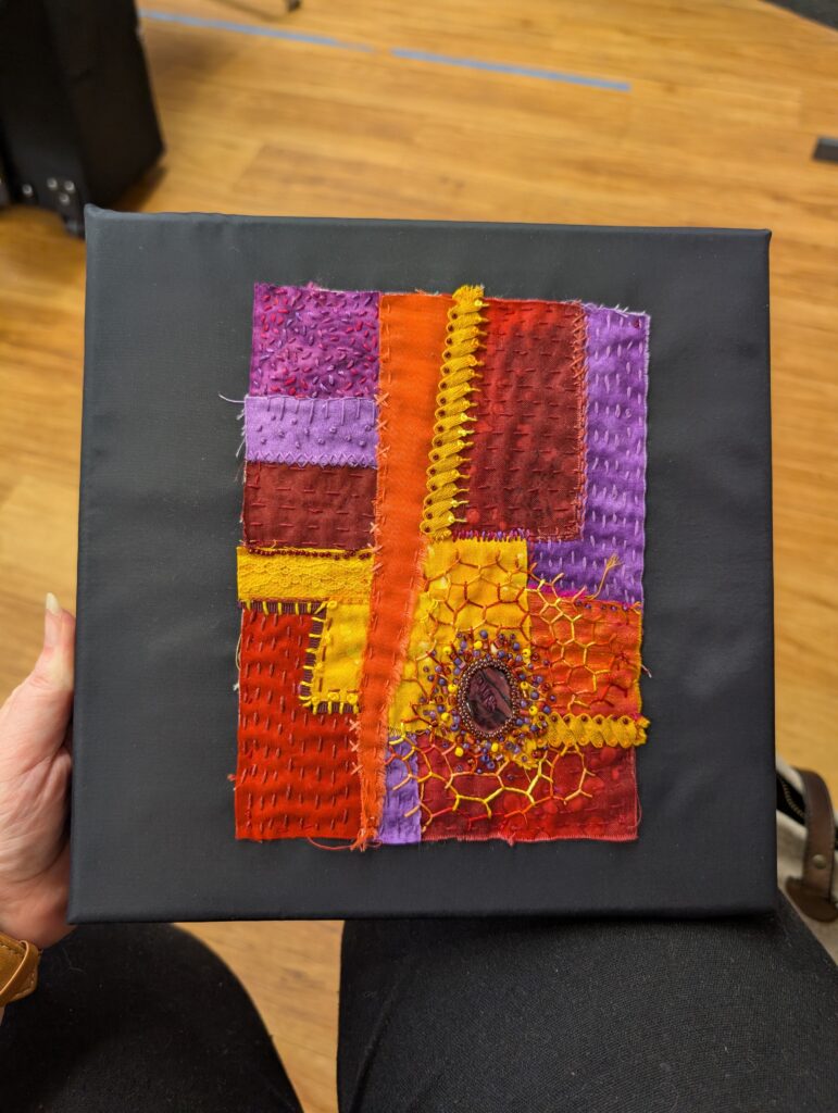 A piece of patchwork fabric mounted on a black fabric stretched frame. The piece has orange, yellow, and purple fabrics embroidered over in matching thread in a variety of stitches. In the lower right there is a beaded oval with a radiating honeycomb stitch pattern.