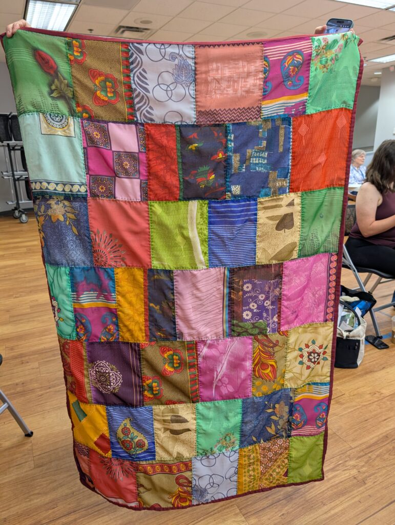 A blanket made up of large squares of printed rayon in pinks, greens, yellows, and blues with an edge bound in maroon. Not technically a quilt because it isn't layered. The fabrics were all salvaged from a rag rug purchased at a discount store.