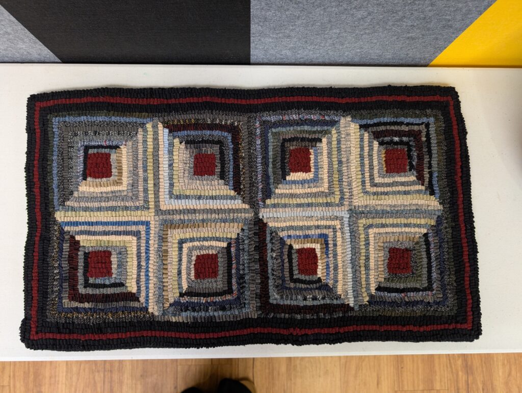 A hand made rug hooked rug done in the classic log cabin quilt pattern. The center squares are dark red, half the blocks diagonally are in dark grey and the other half in light creams and greys. They are arranged so that the light halves form two large lighter diamonds on a darker field.