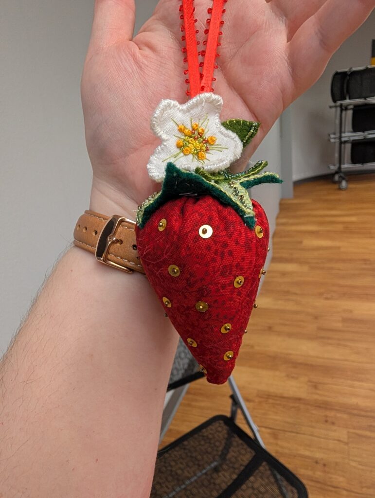 A sweet sewn and stuffed red strawberry Christmas ornament. The strawberry is red and dotted with sequins and a single tiny bead to represent seeds. One the top are green leaves and a white flower with beading and embroidery.