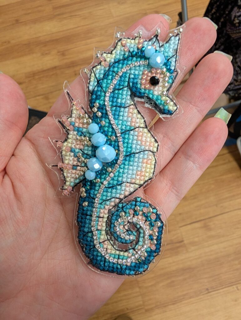 A blue and cream cross stitched sea horse done on a clear plastic form. It is embellished with beads.