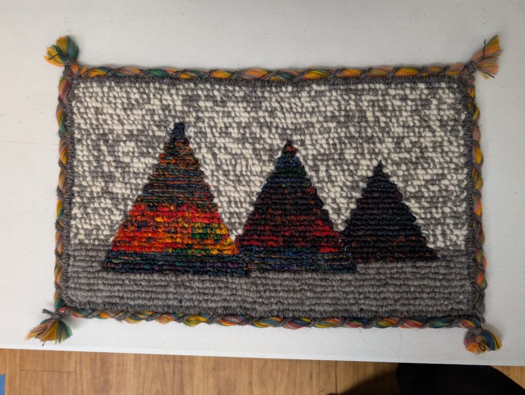 A woven rug depicting a landscape of three trees. The ground is dark grey, the sky is a marled grey and white. Three dark triangles of descending size march across the piece, overlapping slightly. The largest has a wide band of fiery red.
