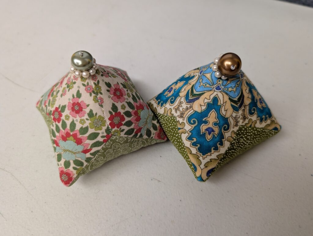 Two pyramidal bean bags. They have been fussy cut and hand sewn together so each face has the same fabric on it. At the pinnacle is a large bead sitting on top of a smaller ring of pearl beads. The one on the left is green and pink florals on a cream background with a green bottom and silver bead. The one on the right is a blue and cream filigree with a green bottom and golden bead.