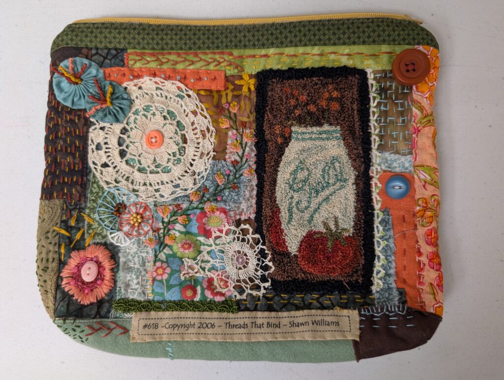 The front side of an embroidered zipper pouch in greens, oranges and blues. It features a small rug hooked depiction of a Ball mason jar, four buttons, two white crochet doilies, and two blue fabric yo-yos. It also has lots of different sashiko stitches in matching and contrasting threads.