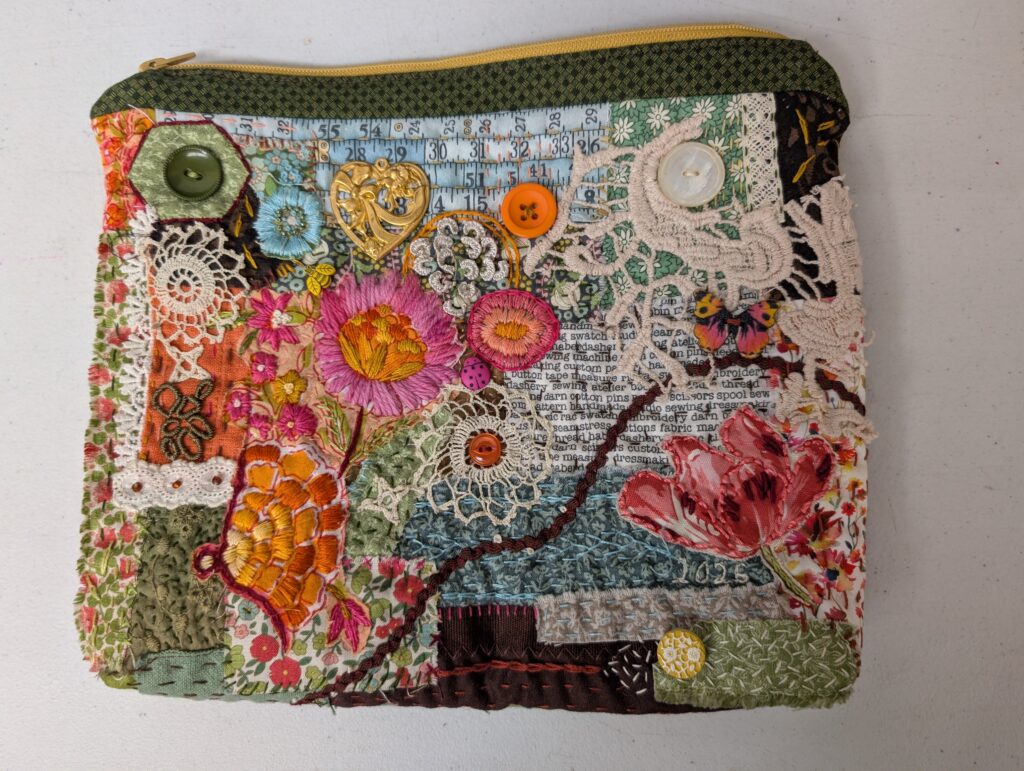 The backside of the heavily embroidered zipper pouch in greens, blues, and oranges. It features five buttons, two doilies, three different kinds of lace, applique flowers, a pink ladybug, an embroidered flowers, and extensive sashiko stitching.