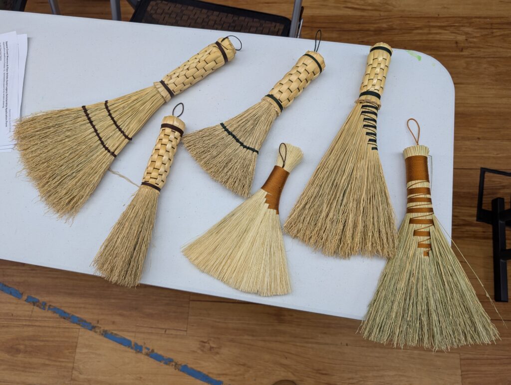 Six hand brooms laid out on a table. Four have woven stalk handles, and two are just wrapped. Two have stitching to spread them out.