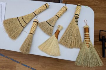 Six hand brooms laid out on a table. Four have woven stalk handles, and two are just wrapped. Two have stitching to spread them out.
