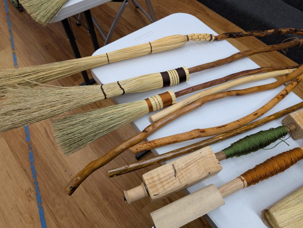 Top to bottom: three hand made cobweb brooms (long thin brooms for cleaning high corners), four broomsticks without straw yet, and two hand made broom spindles, one loaded with green thread, the other with orange.
