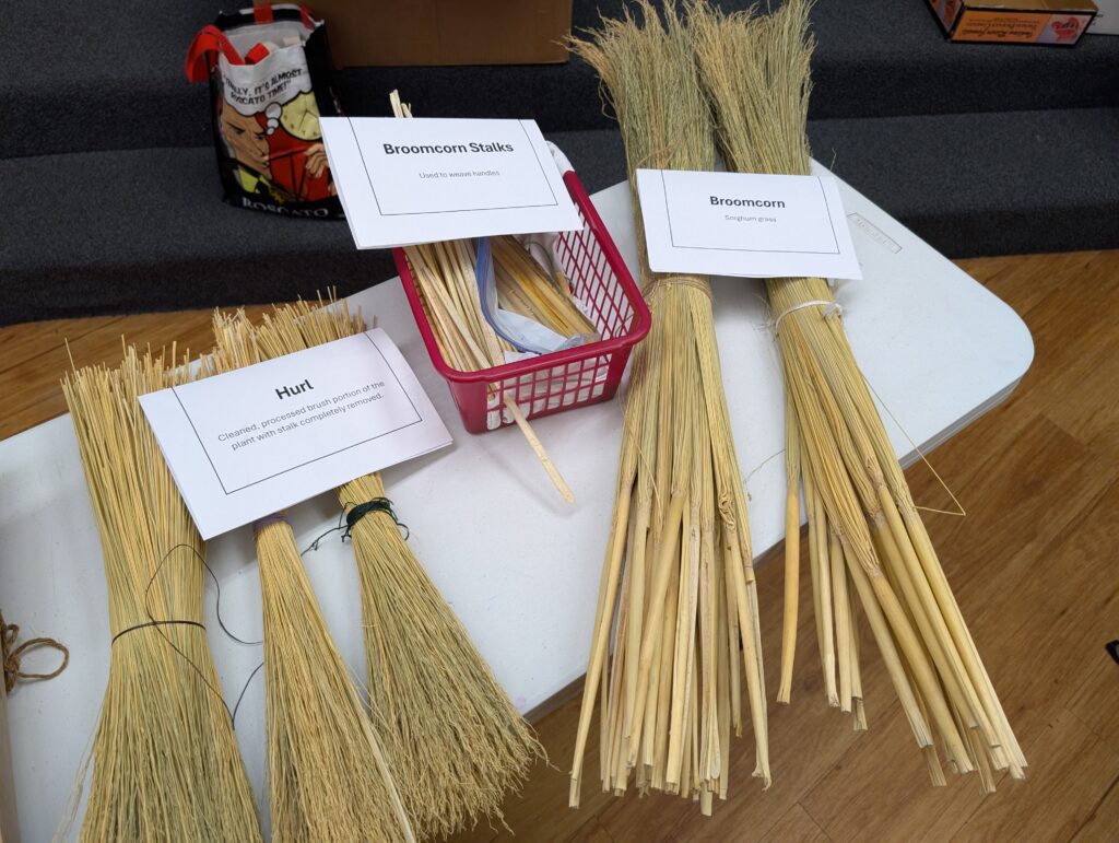 Left to right: three bundles of hurl, a basket of cut broom stalks, and two bunches of raw broomcorn.