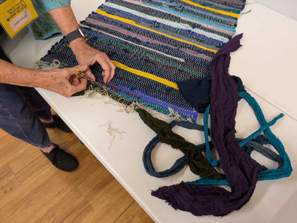 Judy Schwab snipping the warp on a dark colored rag rug in order to harvest the weft from it, which are several dark colored strips of fabric.