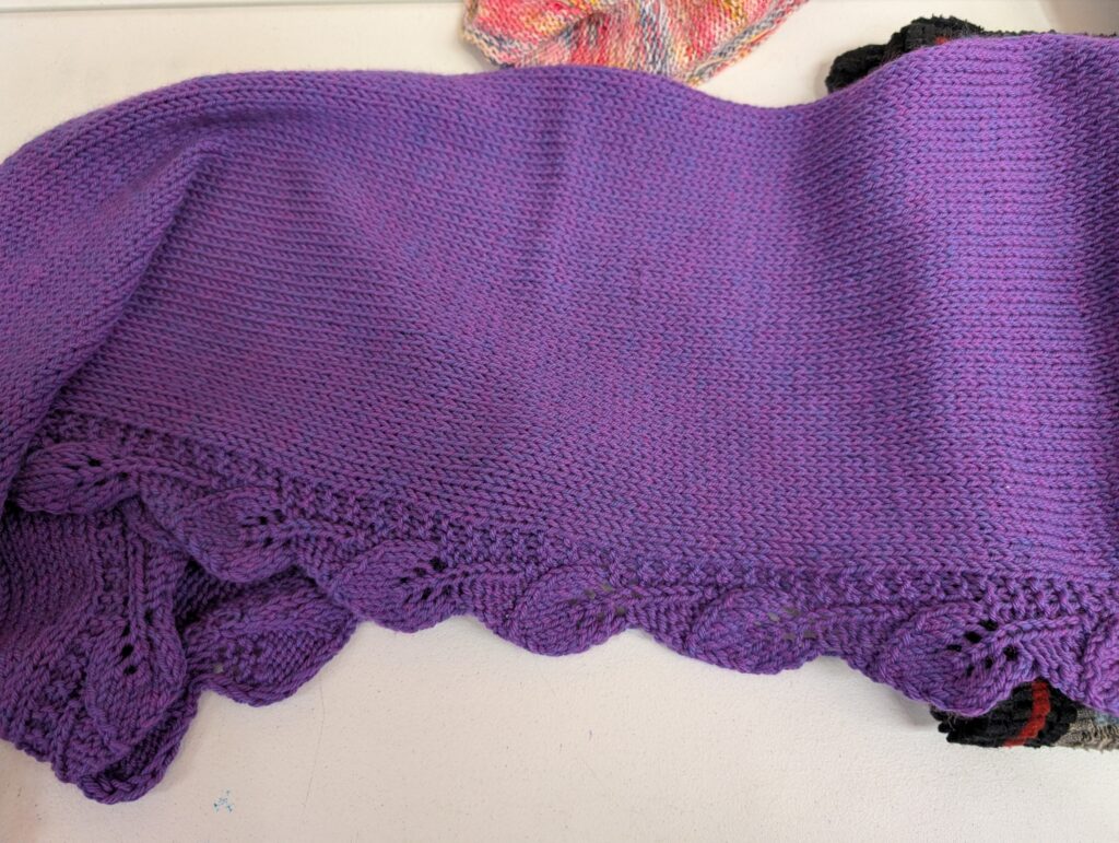 Picture of a purple knit scarf shawl. The body is plain stockinette stitch but the bottom edge features a row of leaves.