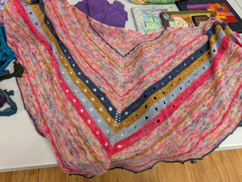 A striped knit triangle shawl in variegated pink yarn. The center features four stripes in dark blue, gold, silver, and pink that have large lace holes.