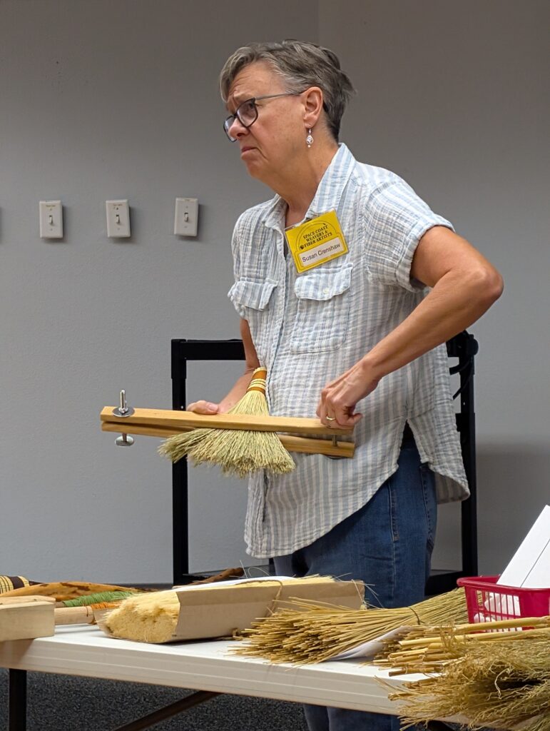 An older woman with short grey hair (Susan Crenshaw) is clamping the end of a broom with a wooden clamp to spread it out for sewing.
