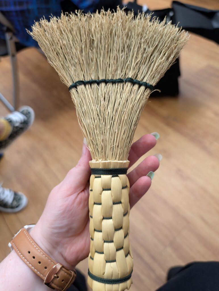 A hand broom with a woven stalk handle. It has a line of dark green thread keeping the corn spread.