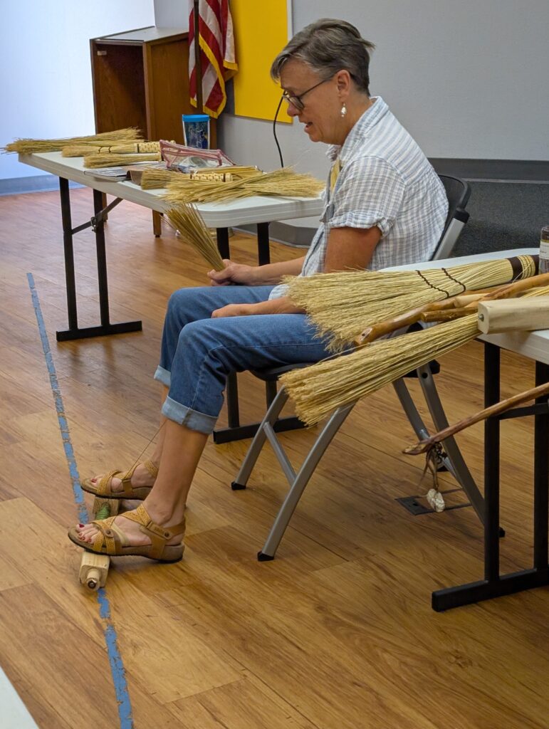 An older woman with short grey hair (Susan Crenshaw) sits in a chair preparing to start wrapping a broom. She holds a bundle of broomcorn in her right hand, he left is holding thread from the spindle. Her feet are braced on the two blocks of the spindle on the floor.