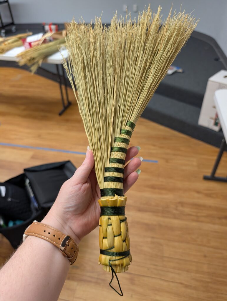 The completed demo hand broom. It is a turkey wing style made of broomcorn, wrapped in green thread with a woven stalk handle.