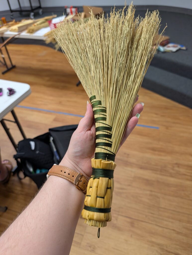 The completed demo hand broom. It is a turkey wing style made of broomcorn, wrapped in green thread with a woven stalk handle.